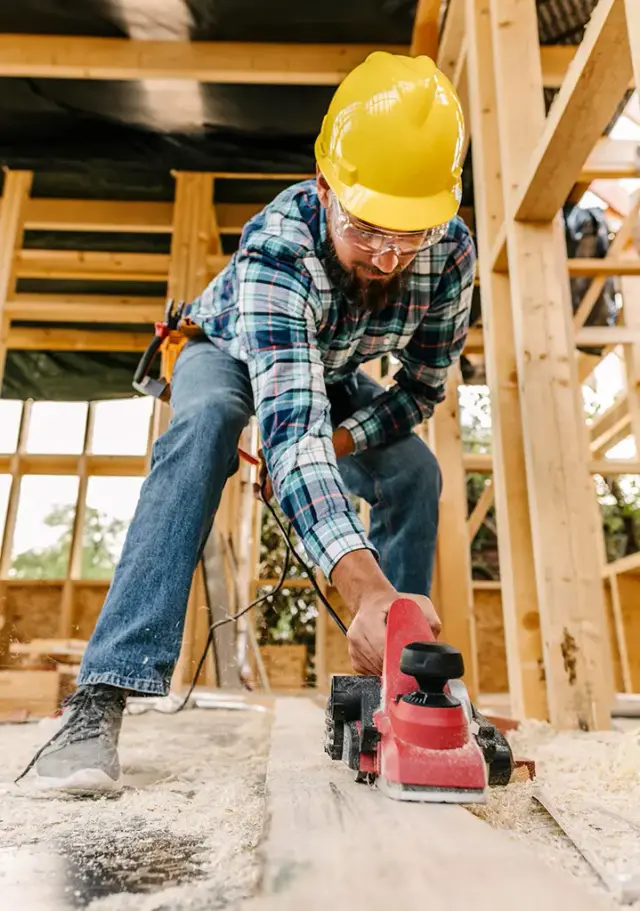 Carpenter wearing a hard hat using an electric hand planer on timber framing